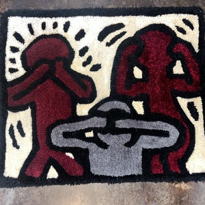 May include: A rug with a design of three figures, two in red and one in gray, with black outlines. The figures are stylized and have their hands covering their eyes. The rug has a white background.