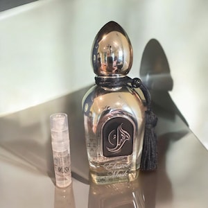 May include: A silver bottle of Elusive Musk perfume with a black tassel. The bottle is sitting on a silver surface next to a clear glass sample vial.