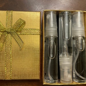 Könnte beinhalten: Eine goldene Geschenkbox mit einer goldenen Schleife, die drei klare Glasflaschen mit Zerstäubern enthält. Die Flaschen sind mit einer klaren Flüssigkeit gefüllt.