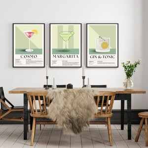 May include: Three framed art prints featuring cocktail illustrations: Cosmo, Margarita, and Gin & Tonic. Each print has a black frame, a light green background, and includes the cocktail name and ingredients. The prints are displayed above a wooden dining table.