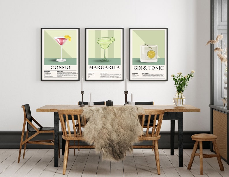 May include: Three framed art prints featuring cocktail illustrations: Cosmo, Margarita, and Gin & Tonic. Each print has a black frame, a light green background, and includes the cocktail name and ingredients. The prints are displayed above a wooden dining table.