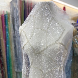 Könnte beinhalten: Weißer, durchsichtiger Stoff mit einem geometrischen Muster aus silbernen Pailletten und Perlen. Der Stoff ist über eine weiße Schaufensterpuppe drapiert.