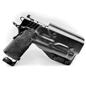 May include: A black semi-automatic pistol partially inserted into a molded black Kydex holster. The firearm features a textured grip, a metallic slide with serrations, and a base plate on the magazine. The holster is secured with two visible screws.