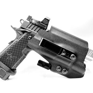 May include: A black handgun with a red dot sight is holstered in a black polymer holster. The holster has multiple screws and a retention strap.