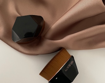 May include: Two black makeup brushes with wooden handles. The brushes have a geometric shape and are made of synthetic bristles. The brush on the left has a flat top and the brush on the right has a rounded top. The brush on the right has the text "MBN BEAUTY" printed on the side.