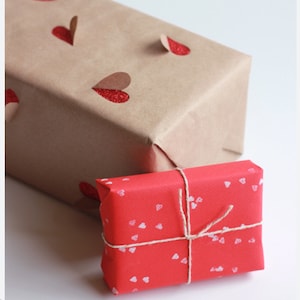 May include: Two wrapped gifts. The larger package is wrapped in brown paper with red glitter heart accents. The smaller gift is wrapped in red paper with white heart patterns and tied with twine.