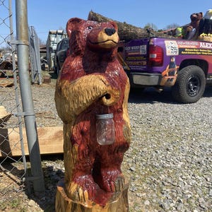 Pode incluir: Estátua de urso de madeira entalhada à mão com acabamento vermelho-castanho, segurando um frasco de vidro. O urso está em pé sobre um tronco de árvore. A estátua está ao ar livre em um dia ensolarado.