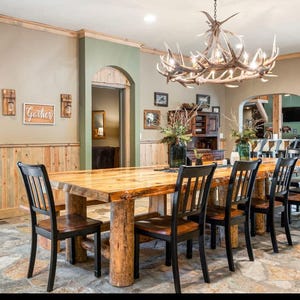 May include: A large rectangular dining table with a natural wood grain surface and thick log legs, surrounded by eight black wooden chairs with slatted backs. Above the table hangs a large chandelier made of antlers with multiple light bulbs.