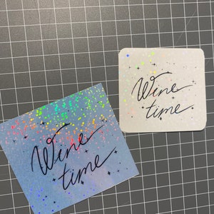 以下が含まれることがあります： 虹色のキラキラした紙2枚に、黒い筆記体で「Wine time」と書かれています。