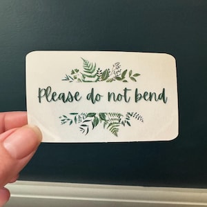 Puede incluir: Una tarjeta blanca con un borde de follaje verde y el texto "Please do not bend" en negro.