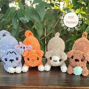 Peut inclure: Quatre jouets en peluche crochetés, un chat violet, un chat orange, un chien brun et un chien beige, sont alignés sur une surface en bois. Les jouets sont tous en position assise, la tête légèrement penchée en avant. Le texte "PDF Pattern" est visible dans un cercle en arrière-plan.