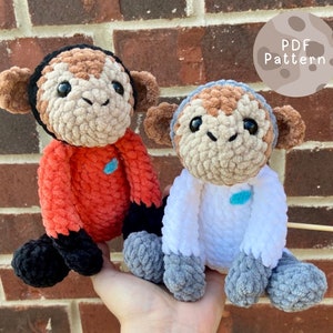May include: Two crocheted monkey plushies, one in orange and one in white, with black and grey accents. The monkeys are sitting on a hand, with a wooden stick behind them. The text "PDF Pattern" is visible in the background.