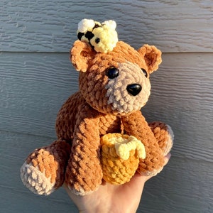 Peut inclure: Un ours en peluche crochet&eacute; marron avec une abeille crochet&eacute; jaune sur la t&ecirc;te. L'ours tient un nid d'abeille crochet&eacute; jaune.