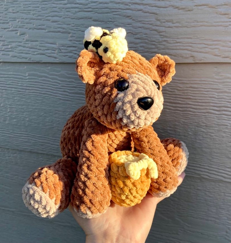 Peut inclure: Un ours en peluche crochet&eacute; marron avec une abeille crochet&eacute; jaune sur la t&ecirc;te. L'ours tient un nid d'abeille crochet&eacute; jaune.