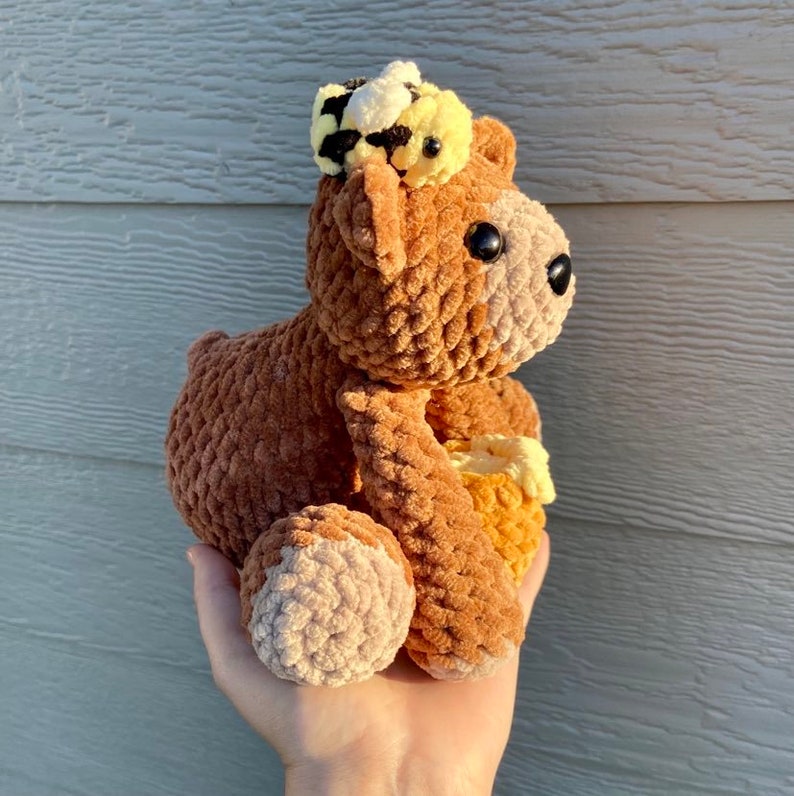 Peut inclure: Un ours en peluche crochet&eacute; marron portant un chapeau d'abeille crochet&eacute; jaune et noir. L'ours tient un nid d'abeille crochet&eacute; jaune.