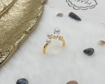 May include: A gold ring adorned with five white pearls and a small, clear gemstone. The ring's design is open and delicate. It is presented on a white, fluffy surface, with a gold leaf and small seashells in the background. The ring is a piece of jewellery.