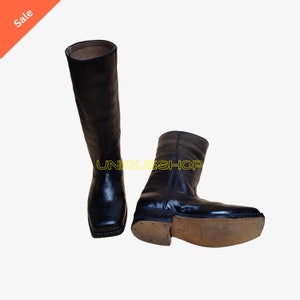 K&ouml;nnte beinhalten: Ein Paar schwarze Lederstiefel mit hohem Schaft und quadratischer Zehenpartie. Ein Stiefel steht aufrecht, w&auml;hrend der andere auf der Seite liegt und eine hellbraune Sohle zeigt. Der Text UNIQUESHOP ist in gelben Buchstaben zu sehen.