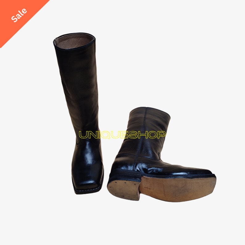 K&ouml;nnte beinhalten: Ein Paar schwarze Lederstiefel mit hohem Schaft und quadratischer Zehenpartie. Ein Stiefel steht aufrecht, w&auml;hrend der andere auf der Seite liegt und eine hellbraune Sohle zeigt. Der Text UNIQUESHOP ist in gelben Buchstaben zu sehen.