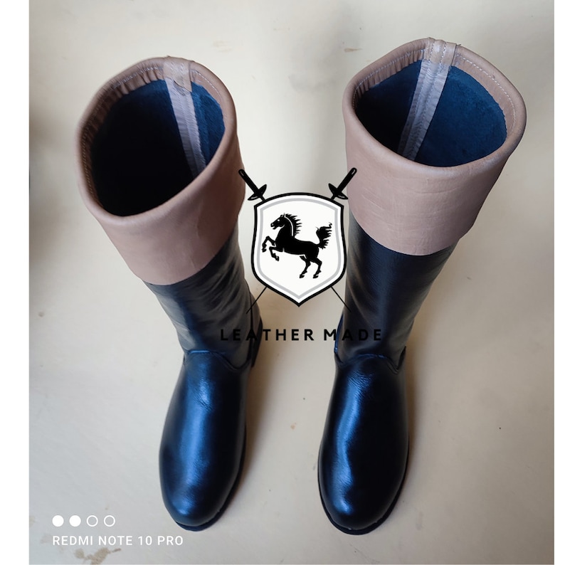May include: A pair of black leather riding boots with tan leather tops. The boots have a pointed toe and a zipper on the inside of the boot. The boots are made by "Leather Made".