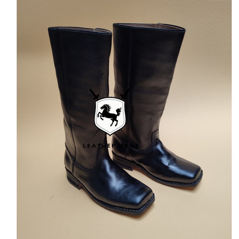 K&ouml;nnte beinhalten: Ein Paar wadenhohe Stiefel aus schwarzem Leder mit quadratischer Zehenpartie und niedrigem Absatz. Ein wei&szlig;es, schildf&ouml;rmiges Logo mit einer schwarzen Pferdesilhouette und zwei gekreuzten Schwertern ist zwischen den Stiefeln mit dem Text LEATHER MADE darunter.