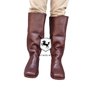 K&ouml;nnte beinhalten: Ein Paar braune, kniehohe Lederstiefel mit quadratischer Spitze. Die Stiefel sind aus glattem Leder gefertigt und haben ein schlichtes Design.