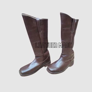 May include: A pair of dark brown leather knee-high boots with a square toe design, low heels, and vertical side pull tabs. The text LEATHER MADE is printed in black outlined capital letters between the two boots against a plain background.