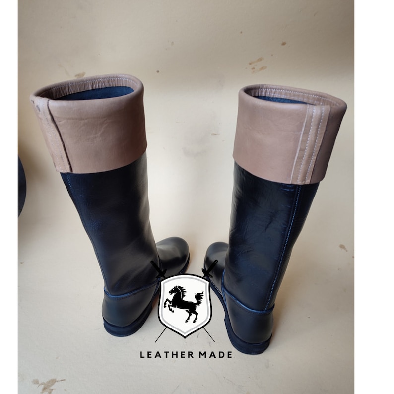 May include: A pair of black leather riding boots with brown leather tops. The boots have a simple design and a rounded toe.