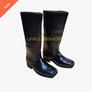 K&ouml;nnte beinhalten: Ein Paar schwarze wadenhohe Stiefel mit quadratischer Zehenpartie und niedrigem Absatz. Die Stiefel verf&uuml;gen &uuml;ber ein strukturiertes Lederobermaterial und eine glatte Oberfl&auml;che im Zehenbereich. Der Text UNIQUESHOPS ist gelb auf die Stiefel gedruckt.