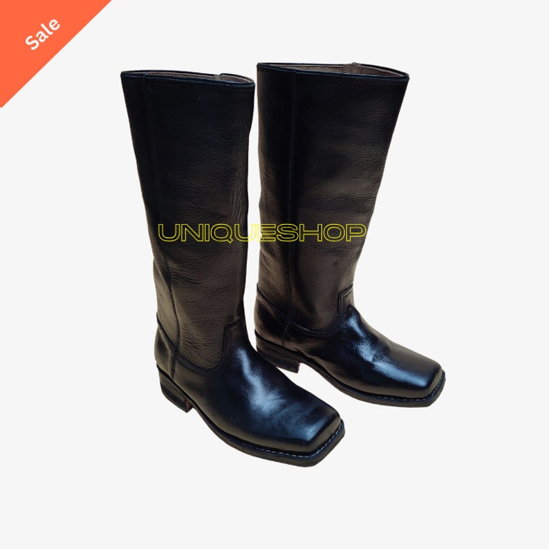 K&ouml;nnte beinhalten: Ein Paar schwarze wadenhohe Stiefel mit quadratischer Zehenpartie und niedrigem Absatz. Die Stiefel verf&uuml;gen &uuml;ber ein strukturiertes Lederobermaterial und eine glatte Oberfl&auml;che im Zehenbereich. Der Text UNIQUESHOPS ist gelb auf die Stiefel gedruckt.