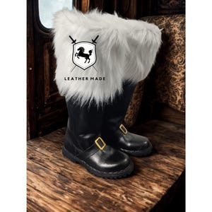 May include: A pair of black leather boots with white faux fur trim. The boots have gold-colored buckles and are displayed on a wooden surface. The boots have the text "LEATHER MADE" on them.