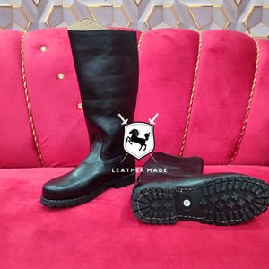 May include: Black leather riding boots with a rugged sole, displayed on a pink velvet surface. The boots are tall, reaching the knee, and feature a classic design. The image includes the text "LEATHER MADE" with a horse logo.