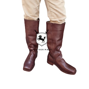 K&ouml;nnte beinhalten: Ein Paar braune, kniehohe Lederstiefel mit quadratischer Spitze. Die Stiefel sind aus glattem Leder gefertigt und haben einen Rei&szlig;verschluss an der Innenseite des Stiefels.
