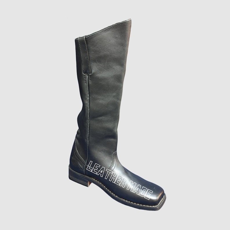 May include: A black knee-high boot with a square toe and a low heel. The side of the boot features the white text LEATHER MADE printed in a bold, sans-serif font. The boot has a smooth finish, visible stitching along the sole, and a tall shaft.
