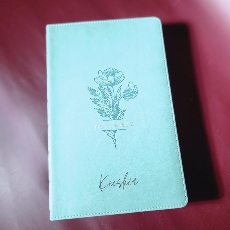 May include: A light teal-colored Bible with a floral design and the words "HOLY BIBLE" in gold lettering. The name "Keeshia" is written in cursive below. The cover has a smooth texture.