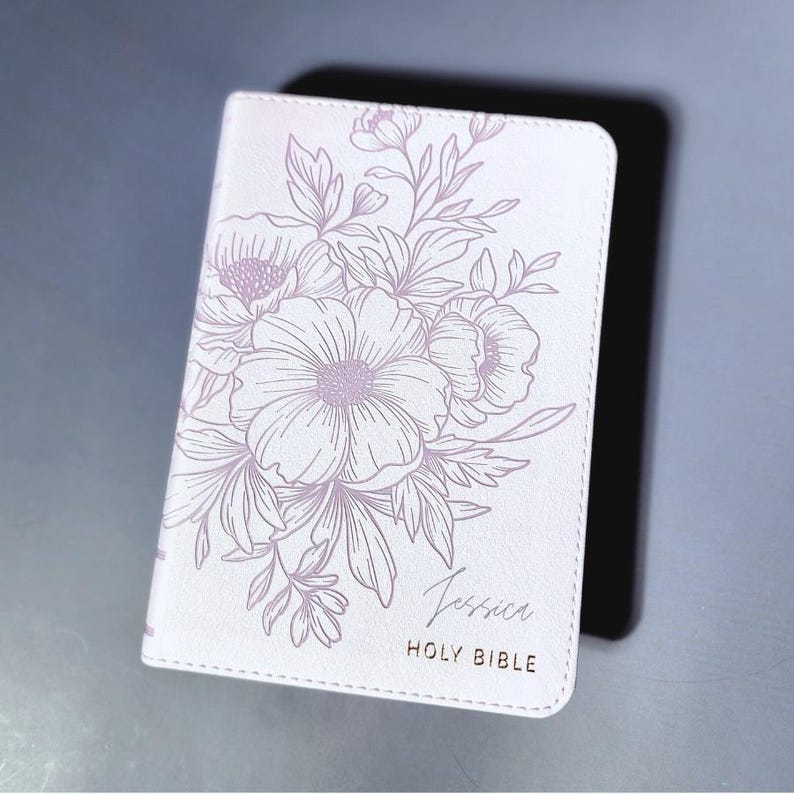 May include: A white Holy Bible with a floral design in light purple. The name "Jessica" is printed in cursive, with "HOLY BIBLE" below it. The cover is resting on a black surface.