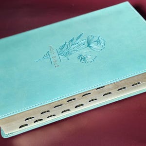 May include: A teal-colored Holy Bible with a floral design embossed on the cover. The words "HOLY BIBLE" are vertically printed on the cover. The book has gold-colored edges and black tabbed pages.