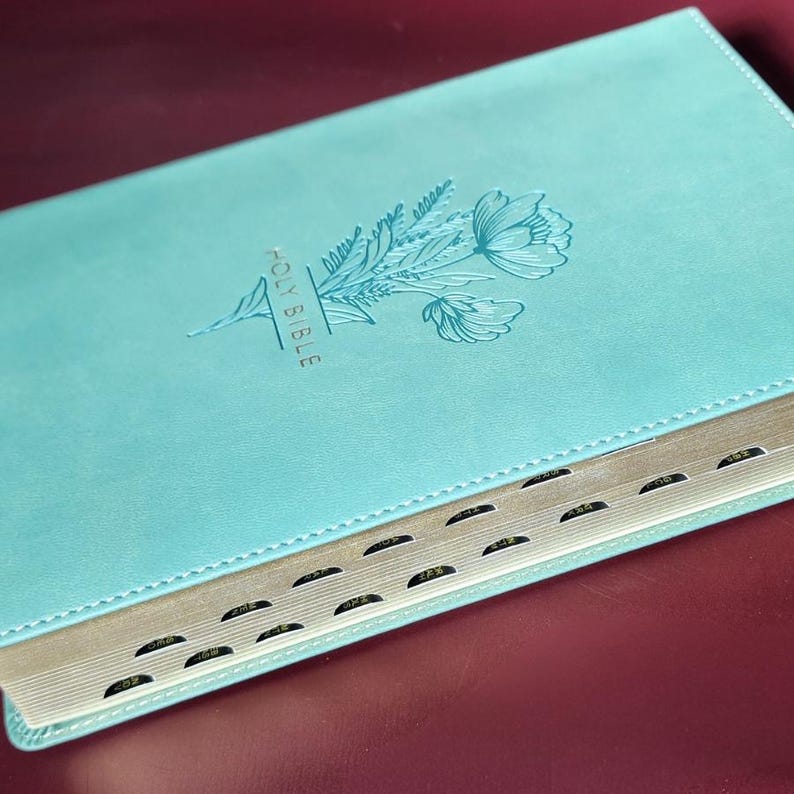 May include: A teal-colored Holy Bible with a floral design embossed on the cover. The words "HOLY BIBLE" are vertically printed on the cover. The book has gold-colored edges and black tabbed pages.
