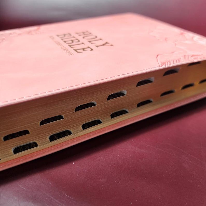 May include: A pink Holy Bible with gold-edged pages. The cover is embossed with the words "HOLY BIBLE" in gold lettering. The pages have thumb index tabs for easy navigation. The book is open, revealing the text within.