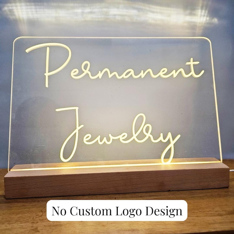 May include: A white acrylic sign with the words "Permanent Jewelry" in a cursive font. The sign is lit from behind and sits on a wooden base. The text "No Custom Logo Design" is printed below the sign.