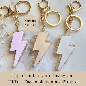 May include: Three gold keychains with lightning bolt shaped resin charms. The charms are in pink, white, and beige with gold flecks. The keychains have a tag that says "Custom NFC Tag".