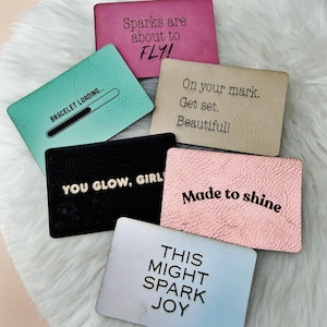 May include: A collection of six rectangular display cards in various colors including magenta, mint green, beige, black, light pink, and white. Each card features unique black text such as Sparks are about to FLY!, BRACELET LOADING..., On your mark. Get set. Beautiful!, YOU GLOW, GIRL!, Made to shine, and THIS MIGHT SPARK JOY.