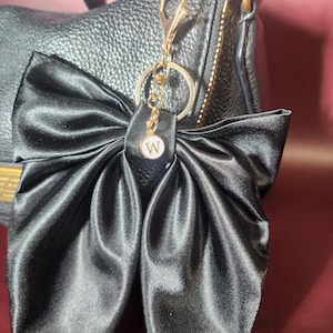 May include: Black satin bow keychain with a gold-colored chain and clasp. The bow is attached to a gold-colored ring and features a small, white circular charm with the letter "W". The keychain is attached to a black leather handbag.