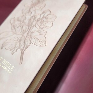 May include: Close-up of a Bible with a floral design embossed on the cover. The cover is a light beige color, and the floral design is a darker beige. The edges of the pages are gold. The text "HOLY BIBLE" and "ES VERSION" are visible.