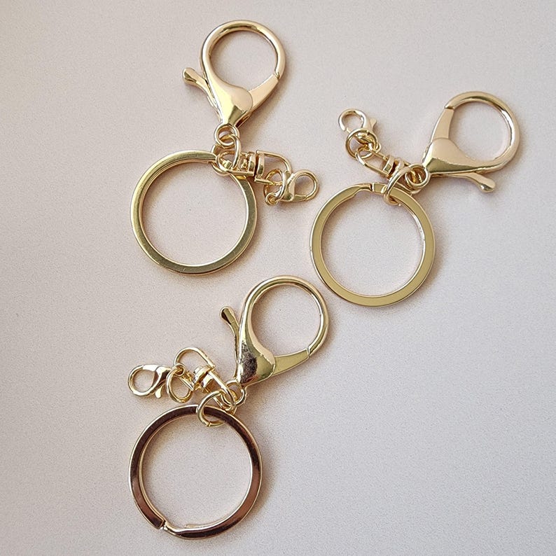 May include: Three gold-colored keychains with lobster claw clasps and large key rings. The keychains are made of metal and have a shiny finish. The key rings are round and open. The clasps are attached to the key rings with small connecting rings.