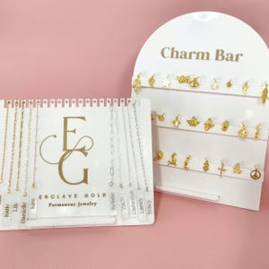 Puede incluir: Una exhibición de collares y dijes de oro y plata sobre soportes blancos. Los soportes tienen el texto "Charm Bar" y "Enclave Gold Permanent Jewelry". Los dijes incluyen hojas, estrellas, abejas y cruces.
