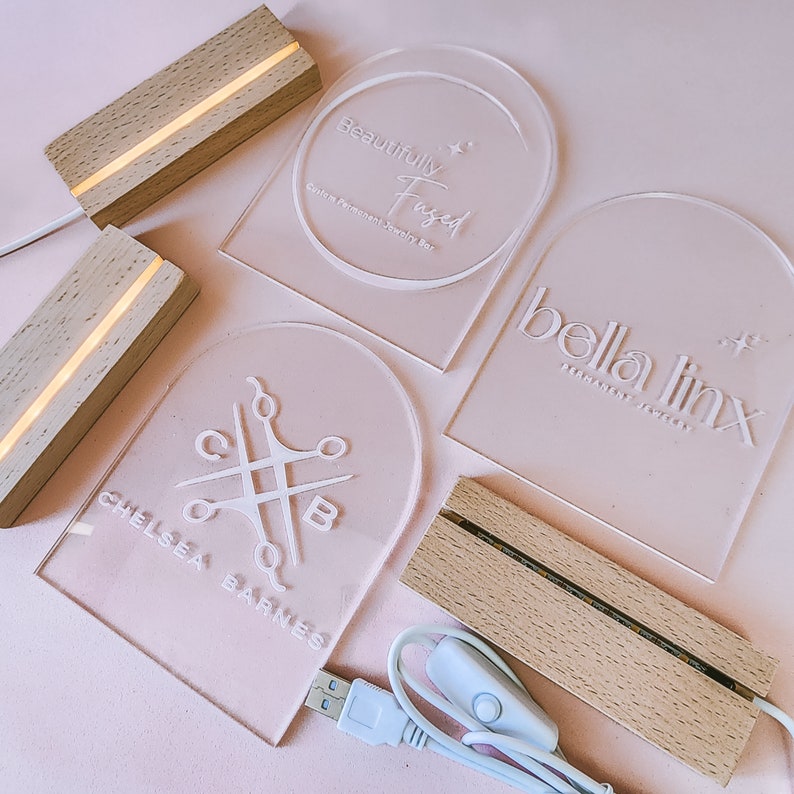 May include: Four acrylic signs with white text and a light source. The signs are on a pink surface. The signs read "Beautifully Fused", "bella linx", "Chelsea Barnes", and a logo with scissors and the letters "C" and "B".