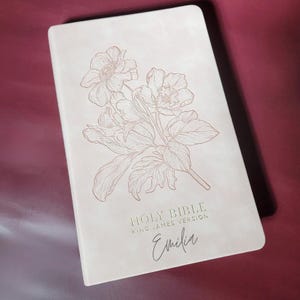 May include: A light beige Holy Bible, King James Version, with a floral design and the name "Emilia" in script. The cover features an embossed illustration of flowers and leaves.
