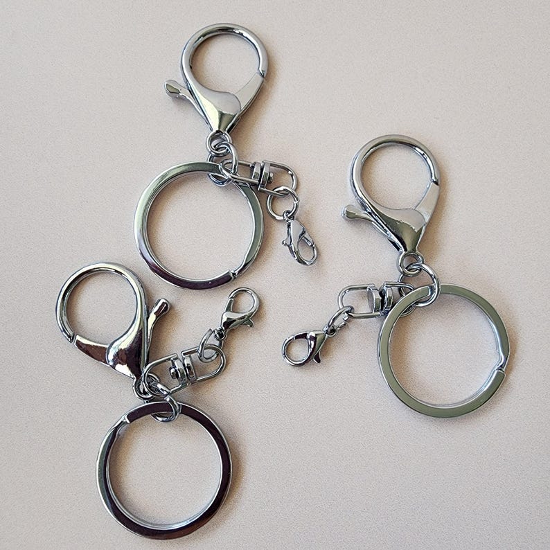 May include: Four silver-colored keychains with lobster claw clasps and split rings. The keychains are made of shiny metal and are arranged on a light beige surface. The clasps are open, ready to attach to a bag or keys.