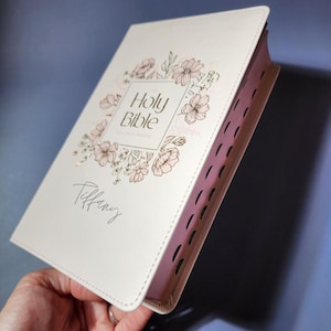 May include: Cream-colored Holy Bible with floral design and the name "Tiffany" in script. The cover features a floral wreath around the words "Holy Bible". The edges of the pages are pink.