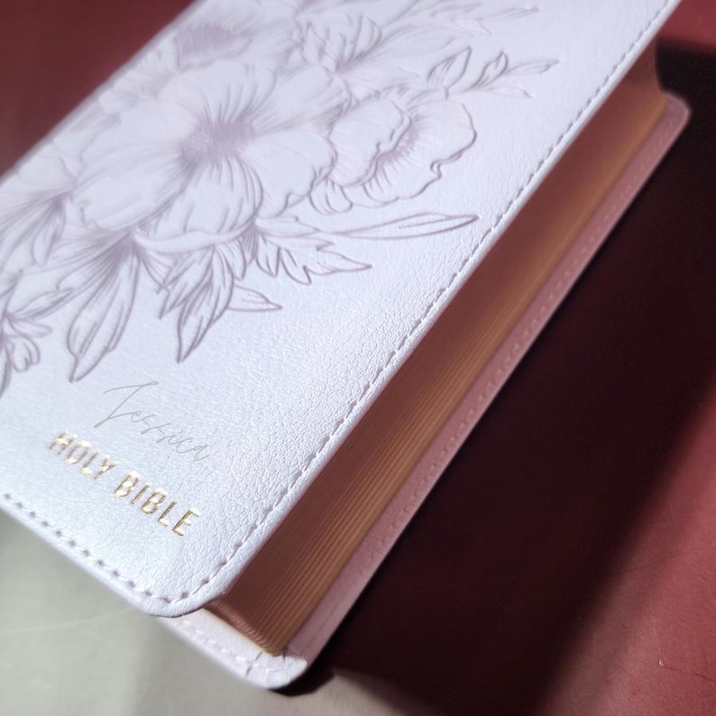 May include: A white Bible with a floral design and the name "Jessica" in script. The words "Holy Bible" are printed in gold. The edges of the pages are a light tan color.